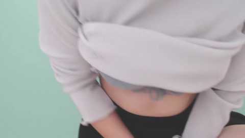 Media: A video of a woman's midsection, wearing a light grey, ribbed crop top that reveals her midriff and black shorts. Her hands are clasped in front, and she has a colorful tattoo on her abdomen. The background is a light green wall.