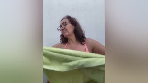 Media: Video of a woman with glasses, wavy brown hair, and a pink top, partially covered by a green towel, leaning against a white textured wall.