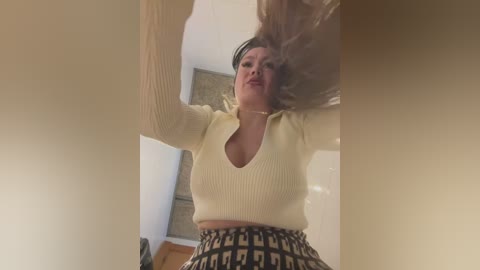 Media: Video of a woman with long brown hair, wearing a cream V-neck sweater and patterned skirt, captured from a low angle, indoors.