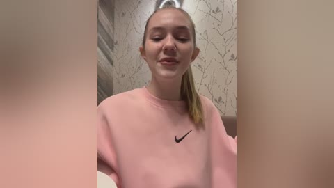 Media: Video of a young woman with fair skin, long blonde hair tied back, wearing a pastel pink Nike sweatshirt, smiling softly, in a bathroom with beige wallpaper and a wooden door.