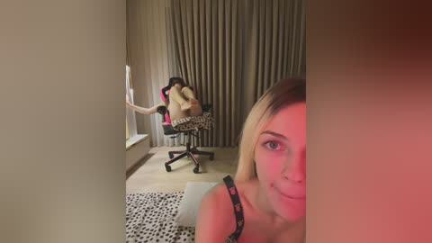 Media: Video of a woman with blonde hair and fair skin, wearing a black tank top, standing in a dimly lit room with beige curtains, leopard-print chair, and a window.