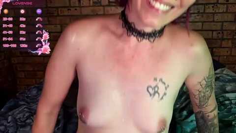 Media: A video of a topless woman with light skin, pink hair, and a black choker, smiling. She has a heart tattoo on her left breast and an elaborate arm tattoo. Background includes a brick wall and a bed with a blue blanket.