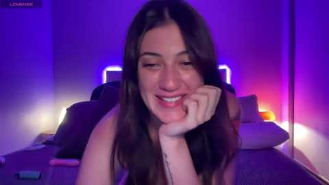 Media: Video of a young woman with long brown hair, smiling, leaning on her hand, in a dimly lit room with purple and blue lighting.