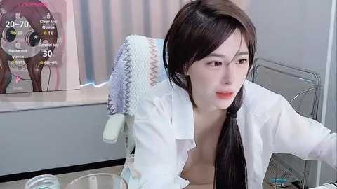 Media: Video of a young, slender East Asian woman with straight black hair in a ponytail, wearing a white lab coat, sitting at a sink in a modern kitchen. A hand towel hangs on a rack behind her.