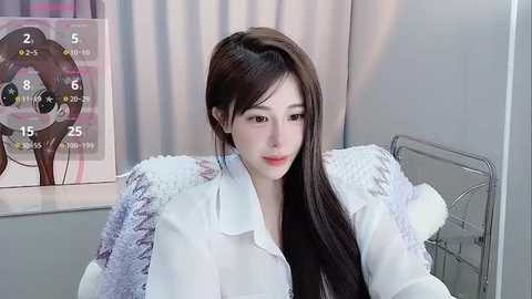 Media: Video of a young Asian woman with long, straight black hair, wearing a white fur-trimmed coat, sitting indoors, with a calendar and a teddy bear visible in the background.