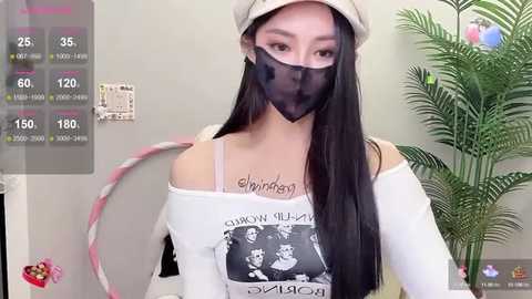 Media: Video of an Asian woman with long black hair, wearing a white off-shoulder top with a graphic, black and white \"Jimi Hendrix\" print, a beige beret, and a black face mask. She stands in a room with a green plant and a pink and white decorative hoop in the background.