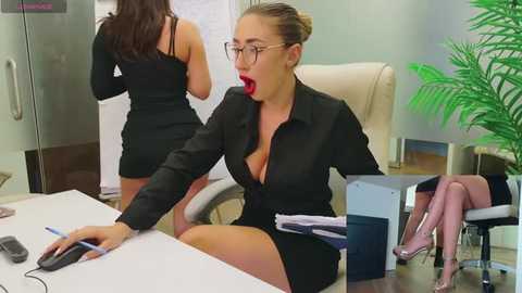 Media: Video of a blonde woman in glasses, wearing a black suit, open shirt, and red lipstick, in an office, holding a phone, with a potted plant in the background.