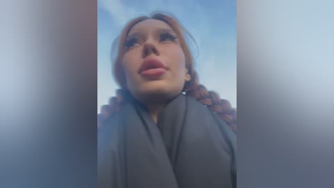 Media: Video of a young woman with fair skin and auburn hair in braids, wearing a gray coat, looking upwards with parted lips. Background is a cloudy sky.