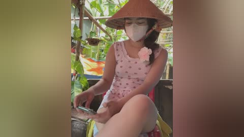 Media: Video of an Asian woman with fair skin, wearing a pink floral dress, a conical hat, and a face mask, sitting on a boat, surrounded by lush greenery.