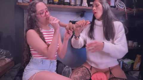 Media: Video of two young women in a messy room; one with glasses, long hair, striped top, blue shorts; other with long hair, white turtleneck, lace thigh-highs, sitting on a pink dildo.