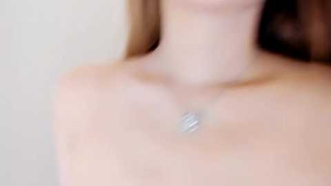 Media: Video of a fair-skinned woman with long brown hair, wearing a delicate silver necklace with a rectangular pendant. The background is blurred, focusing on her neck and shoulders.