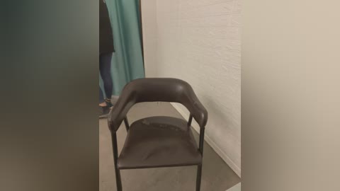 Media: Video of a black, modern, metal-framed chair with a cushioned seat and backrest, positioned in a dimly lit, sparse room with white walls and a teal curtain.