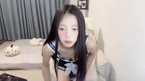 Media: A video of an East Asian woman with long black hair, dressed in a sailor schoolgirl outfit, leaning against a wall in a minimalistic bedroom with light-colored furnishings.