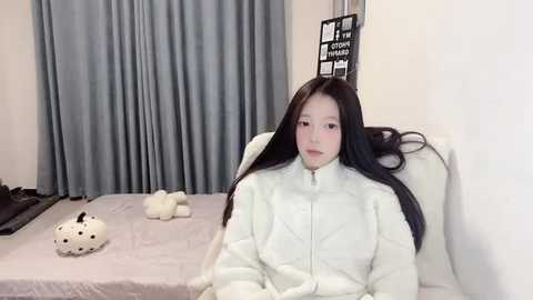 Media: A video of a young East Asian woman with long black hair, wearing a white puffy jacket, seated on a white bed with gray curtains and a stuffed animal, a black shelf with books in the background.