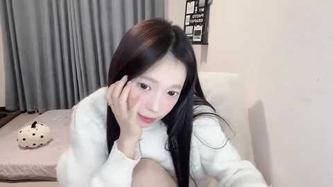 Media: Video of an Asian woman with long black hair, fair skin, wearing a white jacket, lying on a bed with a grey blanket and a stuffed dog.