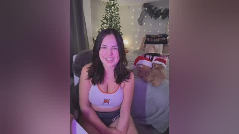 Media: Video of a smiling, light-skinned woman with shoulder-length dark hair in a Hooters tank top, sitting in a cozy, festive room with a decorated Christmas tree and stuffed animals.