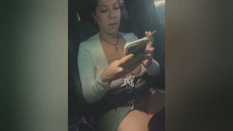 Media: A video of a young woman with light skin and dark hair, wearing a white cardigan and green shorts, sitting in a dimly lit car, playing with her smartphone.