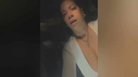 Media: A video shows a woman with light brown skin, wearing a white dress with a plunging neckline, looking distressed. She's standing in a dimly lit room, with blurred background objects.