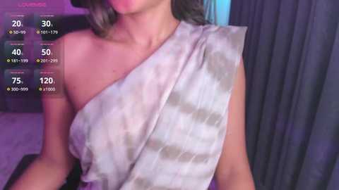 Media: Video of a woman in a gray and white tie-dye one-shoulder dress, with a background of purple and blue curtains.