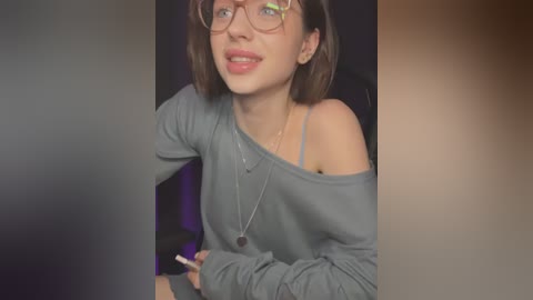 Media: Video of a young woman with short brown hair, glasses, smiling, wearing a gray off-shoulder sweater, holding a brush, indoors with blurred background.