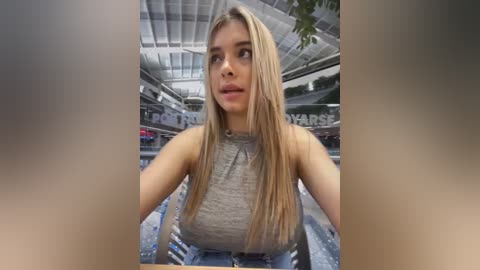 Media: Video of a blonde woman with long hair, wearing a gray sleeveless top, sitting in a car with a blue interior, inside a modern, glass-roofed garage.