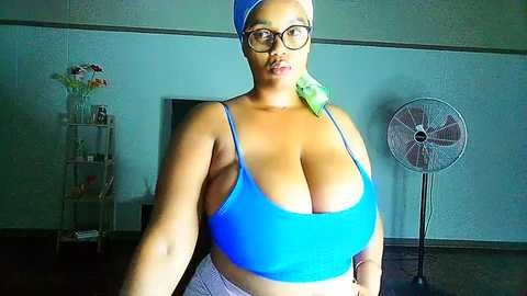 Media: Video of a voluptuous, dark-skinned woman with large breasts, wearing a blue sports bra and headscarf, standing in a dimly lit room with a white fan and flower vase in the background.