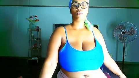 Media: Video of a smiling, plus-sized Black woman with short hair and glasses, wearing a blue sports bra and pink shorts, standing in a modern room with a fan, shelf, and potted plants in the background.