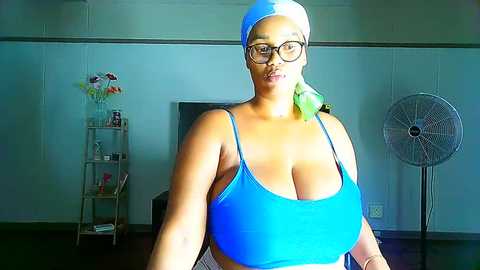 Media: Video of a plus-size, dark-skinned woman with a large bust in a blue sports bra and glasses, standing in a sparsely decorated room with a white fan and a shelf with flowers and books.