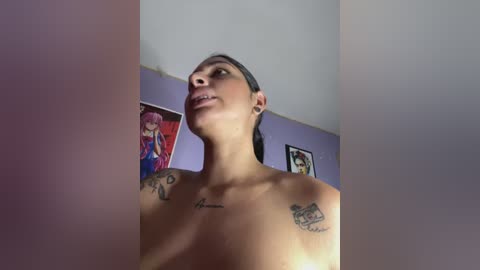 Video of a topless woman with dark skin and dark hair, tattoos on her chest and shoulders, standing in a room with purple walls and framed anime posters.