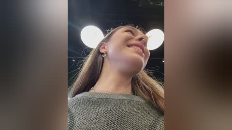 Media: Video of a young woman with straight, light brown hair, wearing a gray sweater, smiling, with two large studio lights in the background, capturing a joyful, candid moment.