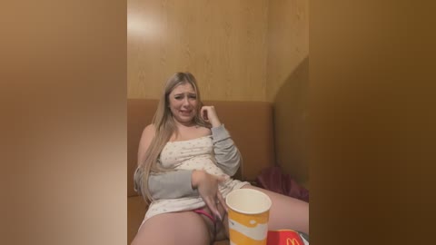 Media: Video of a young Caucasian woman with long blonde hair, wearing a gray tank top and red shorts, sitting on a brown couch, holding a McDonald's cup.