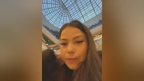 Media: Video of a young woman with light brown hair and light skin, wearing a nose ring, standing indoors near a large, glass-domed ceiling. Background includes shopping mall stores and a busy crowd.