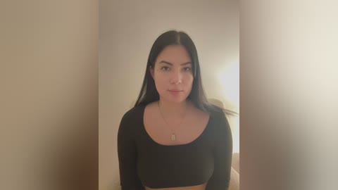 Media: Video of a young Latina woman with long black hair, wearing a black long-sleeve crop top, standing in a dimly lit room with beige walls and a wooden door in the background.