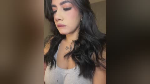 Media: Video of a young woman with long, wavy black hair, wearing a gray tank top, makeup featuring pink eyeshadow and dark eyeliner, and a tattoo on her chest.