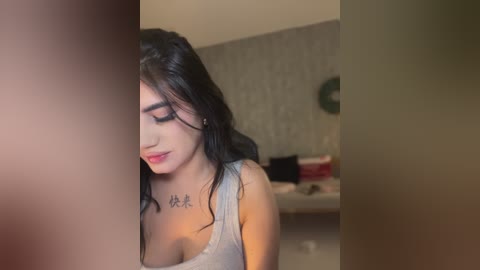 Media: A video shows a young woman with long black hair and fair skin, wearing a gray tank top, leaning against a doorway. She has a tattoo on her chest. The background is a dimly lit bedroom with a bed and patterned wallpaper.