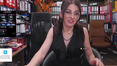 Media: Video of a woman with long brown hair, wearing glasses and a black sleeveless top, sitting at a desk in a cluttered office with colorful bookshelves and a computer screen displaying financial data.