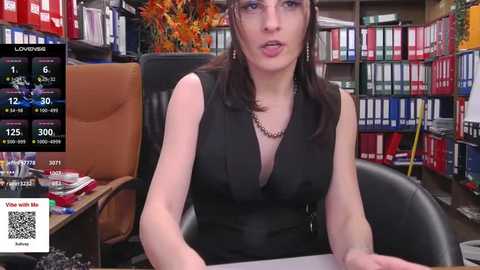 Media: A video of a woman with shoulder-length dark hair, wearing a black sleeveless top, sitting at a desk in an office filled with bookshelves and colorful binders.