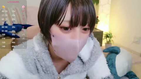 Media: A young Asian woman with short dark hair, wearing a fluffy white and grey robe, has a white surgical mask covering her mouth and nose. She's indoors, with a blurred background featuring a lamp and a potted plant.