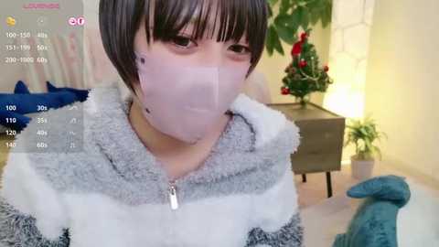 Media: A video of an Asian woman with short dark hair, wearing a gray and white striped pajama top, a white face mask, and a blue furry dinosaur plush. She's in a brightly lit living room with a Christmas tree and potted plants.