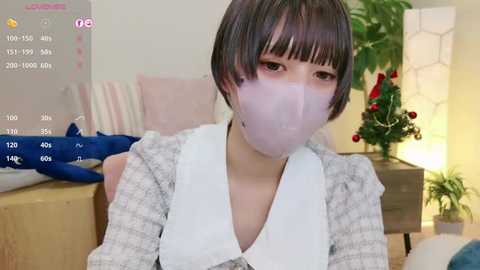 Media: Video of a woman with short black hair, wearing a white mask, gray checkered dress, and seated in a cozy room with a Christmas tree and a lamp.