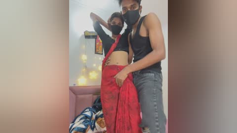 Media: Video of two South Asian individuals in casual attire, standing close in a dimly lit room with string lights. One wears a black mask, black crop top, and red sari, while the other dons a black tank top and jeans.