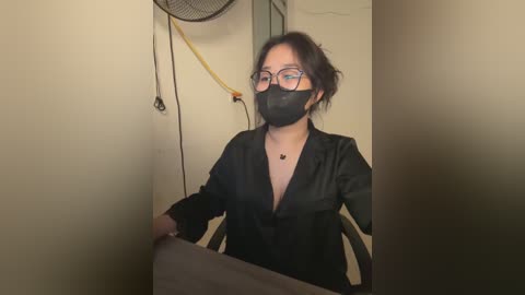 Media: Video of an Asian woman with short dark hair, wearing glasses, a black mask, and a black satin blouse, sitting on a chair in a dimly lit room with beige walls.