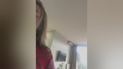 A video of a woman with long, blonde hair partially hidden by a wall, wearing a red Spider-Man shirt, standing in a modern, light-colored room with a TV and window.