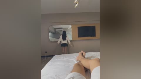 Media: Video of a woman in a white blouse and black skirt, facing a mirror in a modern, minimalist bedroom, with a man in white pajama bottoms in the foreground.