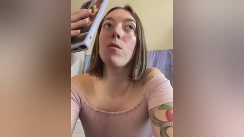 Media: Video of a Caucasian woman with straight, shoulder-length brown hair, wearing a light pink off-shoulder top, taking a selfie in a bathroom. She has a tattoo on her right arm.