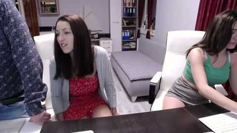 Media: Video of a young brunette woman in a red dress, seated on a desk, flanked by a man in a patterned shirt and another woman in a green tank top. Background shows a modern office with a bookshelf.