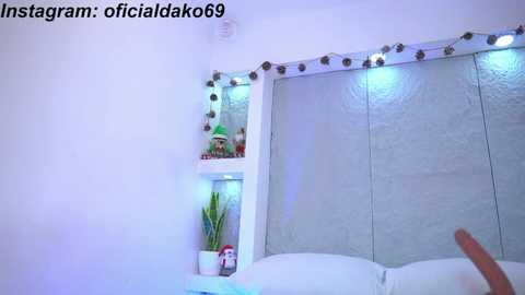 Media: Video of a modern bedroom with a white bed, white walls, and a large window with a white shutter. The room is adorned with string lights and elf-themed decorations, including a small elf figurine.