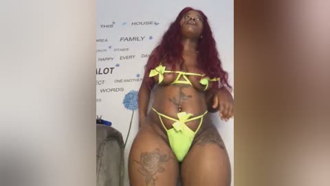 Media: Video of a voluptuous, dark-skinned woman with long, wavy red hair wearing neon green lingerie with strappy details and large floral tattoos on her thighs. Background features a white wall with text promoting \"FAMILY.\