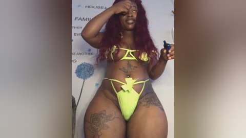 Media: Video of a dark-skinned woman with long, wavy red hair, wearing a neon yellow thong bikini, with tattoos on her thighs and stomach, posing in front of a white wall with text.