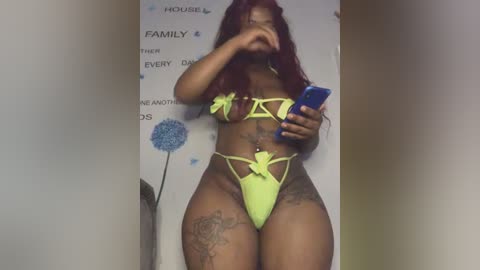 Media: Video of a curvaceous, light-skinned woman with long red hair, wearing neon yellow lingerie with floral accents, taking a mirror selfie. Background features a white wall with family-themed decor.